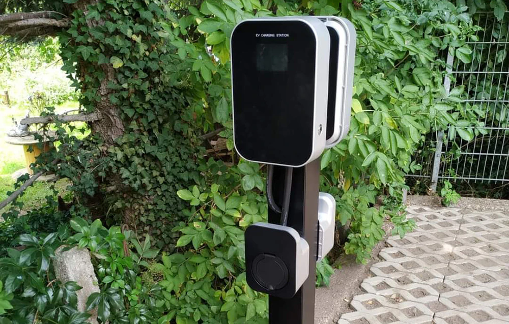 O stație de încărcare pentru vehicule electrice independentă, montată pe un stâlp în grădină, lângă niște plante.