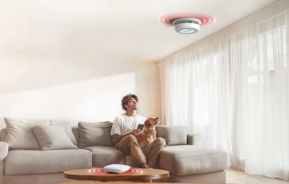 Un bărbat cu un câine pe canapea privește un detector de fum montat pe tavan care comunică cu un gateway Wi-Fi