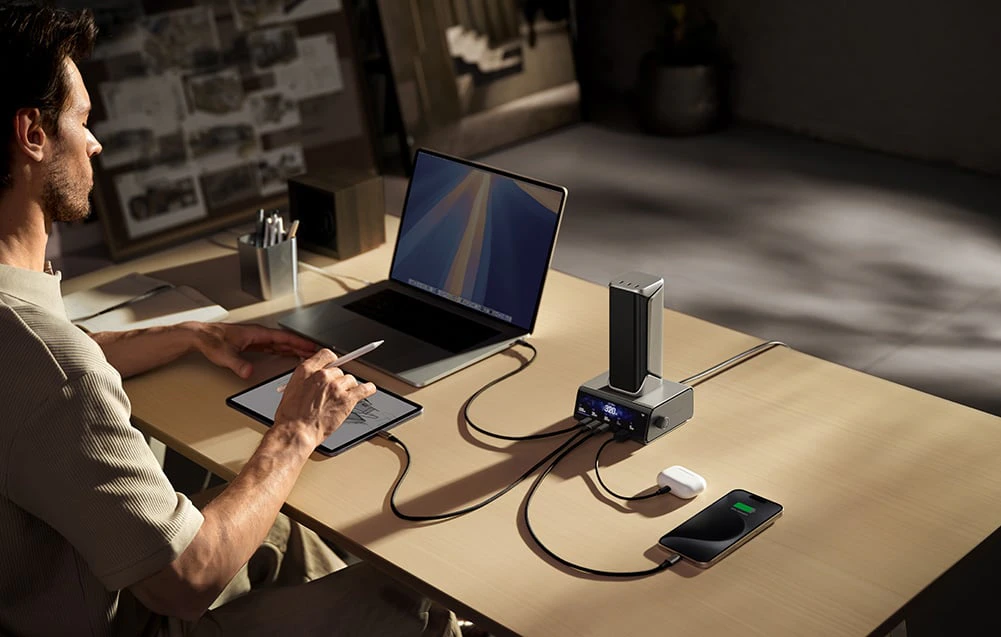 Un bărbat stă la un birou și desenează pe o tabletă. Lângă el sunt un laptop, un smartphone și căști conectate la un powerbank cu stație.