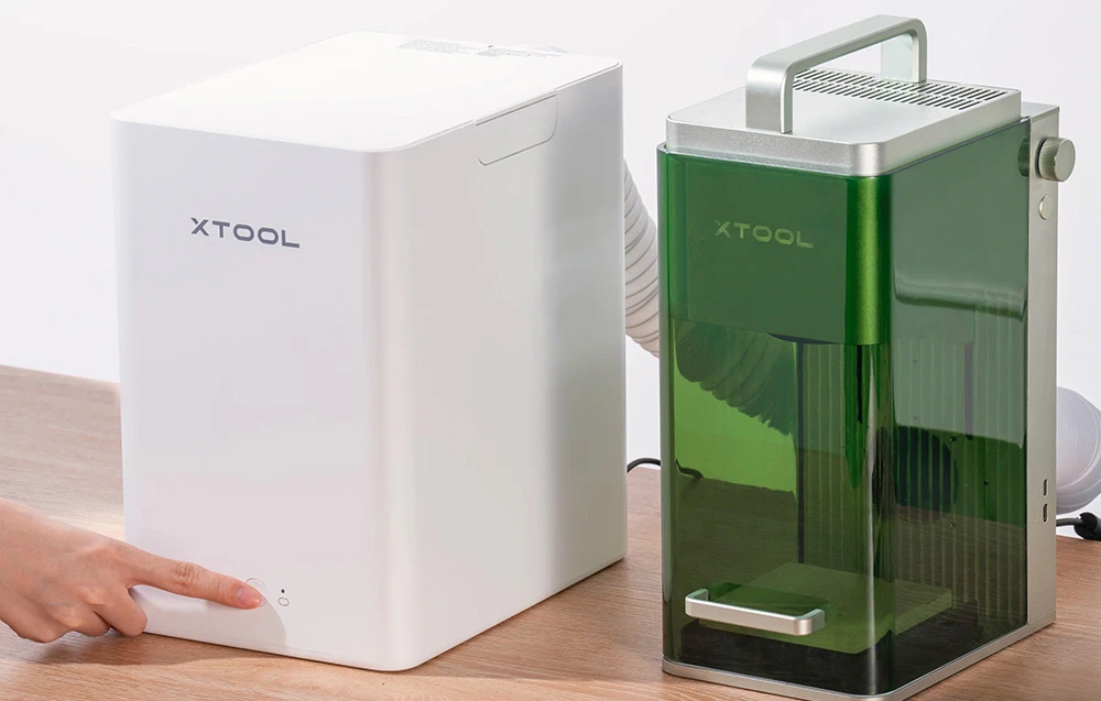 Un deget care activează aspiratorul de fum xTool alb, conectat printr-un tub la un tăietor cu laser verde așezat pe un birou.