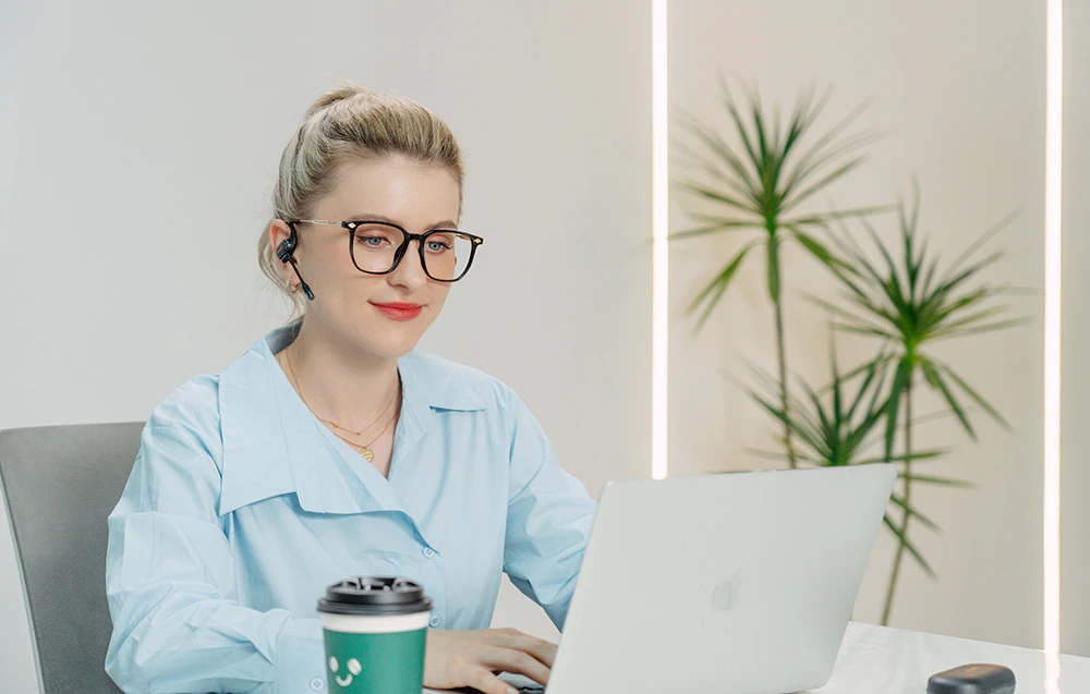 Femeie cu ochelari care lucrează la un laptop, purtând căști in-ear negre cu microfon instalat