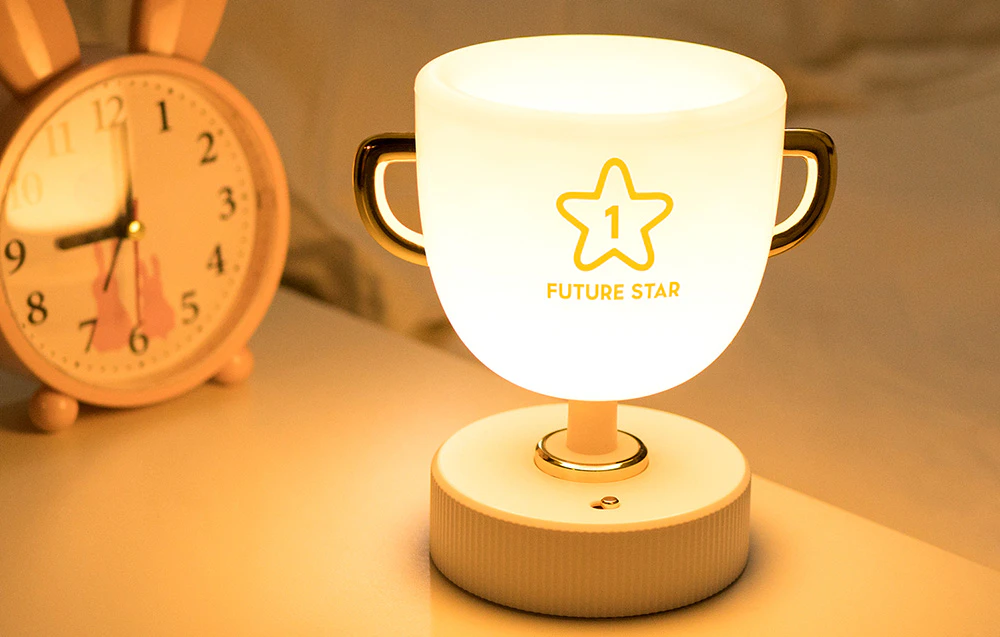 Lumină de noapte pentru copii SuperFire W-006 Champion Light strălucind cu lumină albă. Dispozitivul este așezat pe un dulăpior lângă un ceas deșteptător cu urechi de iepuraș.
