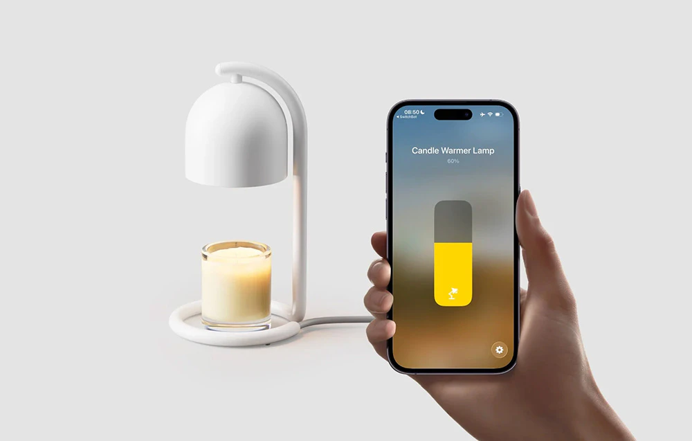 O mână care ține un smartphone cu aplicația SwitchBot, care afișează un glisor setat la 60%, cu un încălzitor de lumânări alb și o lumânare parfumată într-un recipient de sticlă în fundal.