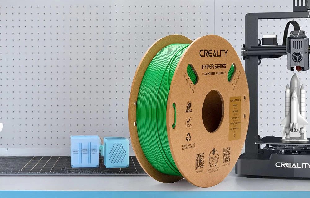 Bobină cu filament verde lângă o imprimantă 3D și modele albastre