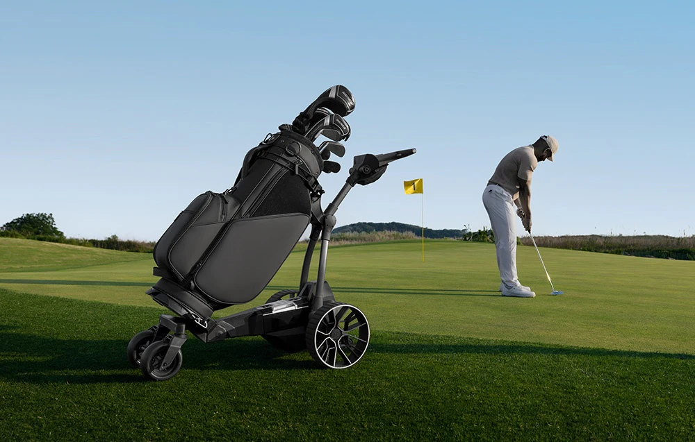 O mașinuță de golf automată cu o geantă stă pe green în timp ce un jucător de golf lovește mingea în gaură