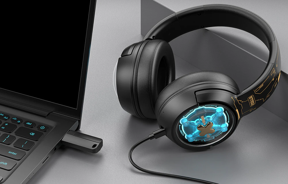 Căști de gaming Onikuma așezate pe un birou lângă un laptop cu un adaptor USB conectat