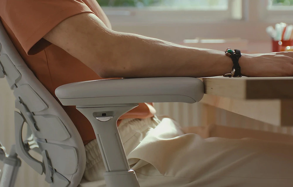 O persoană stă așezată într-un scaun ergonomic de culoare deschisă, sprijinindu-și antebrațul pe cotieră, lângă un birou din lemn