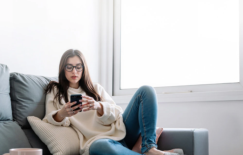 O femeie cu ochelari stă pe o canapea ținând un smartphone