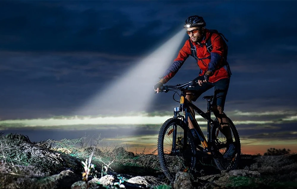 Un bărbat care poartă cască și ochelari merge cu bicicleta noaptea, luminându-și drumul cu lanterna frontală.
