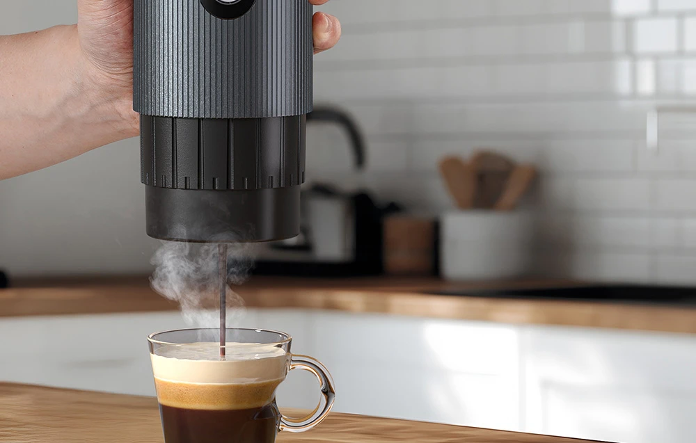 O mână care ține o mașină de espresso de culoare gri închis, din care cafeaua curge într-o ceașcă