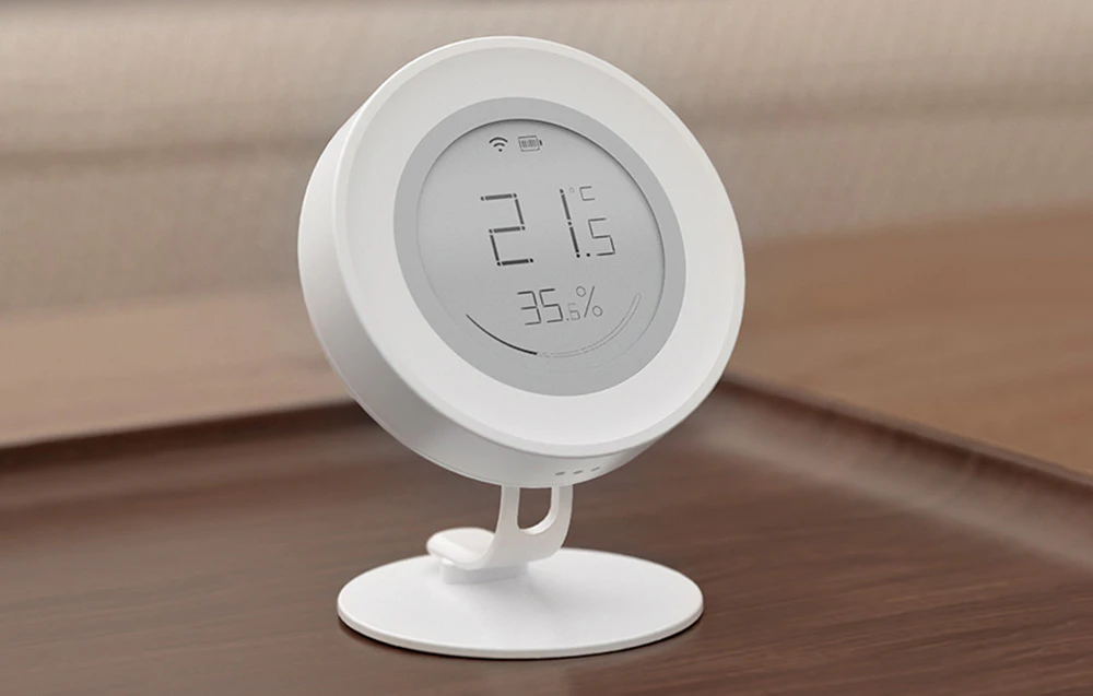 Senzor rotund alb pe un suport care indică temperatura de 21,5°C și umiditatea de 35,6%, așezat pe o masă de lemn