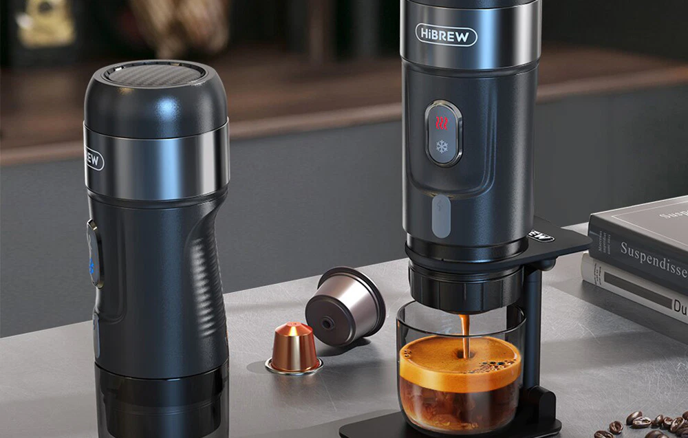 Două aparate de cafea negre stau pe blatul de bucătărie lângă capsule de cafea, un pahar cu cafea și boabe de cafea.