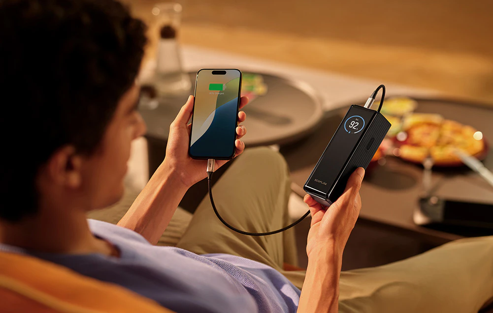 Un bărbat ține telefonul conectat la un powerbank negru deasupra mesei