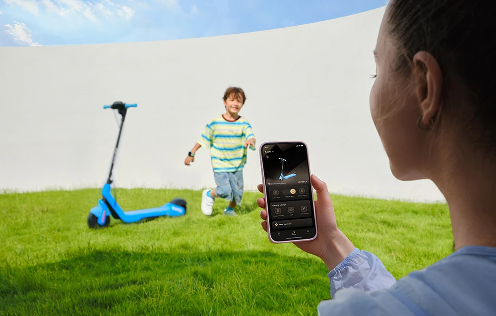 O femeie folosește aplicația de pe smartphone-ul ei, în timp ce un copil aleargă alături de o trotinetă parcată pe iarbă
