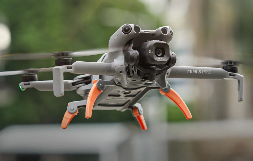 Dronă DJI Mini 5 Pro în zbor, cu picioarele portocalii desfăcute, plutind în aer