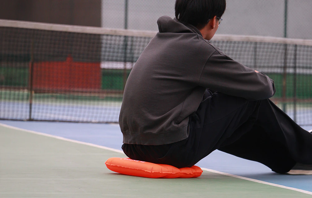 O persoană stă pe o pernă gonflabilă portocalie pe un teren de tenis, sprijinindu-se cu spatele de fileu