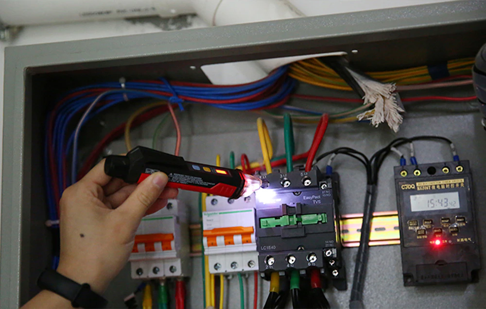 O mână care ține un senzor de tensiune și verifică componentele dintr-un tablou electric cu cabluri și un contor digital.