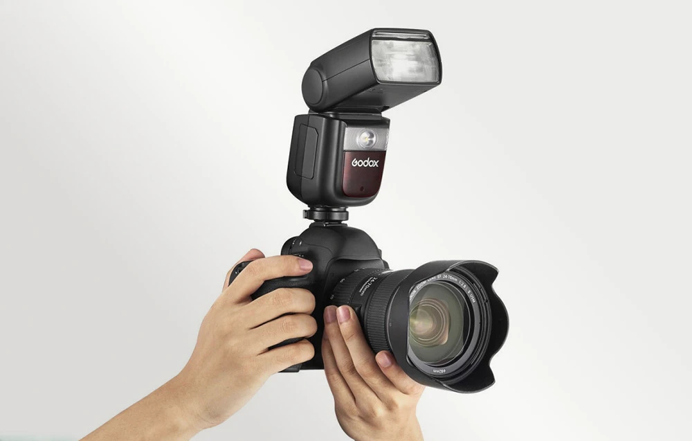 Aparat foto DSLR cu obiectiv și un bliț Godox V860III montat, cu LED-ul aprins, ținut în mâini