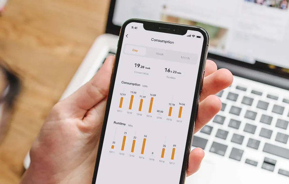 O mână ține un smartphone cu aplicația Consum, care afișează grafice ale consumului de energie electrică și ale timpului de funcționare.