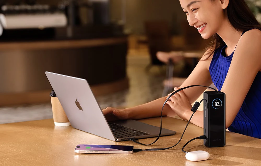 O femeie zâmbitoare stă la o masă cu un laptop deschis și o cană de cafea. Lângă ea se află un smartphone și căști conectate la un powerbank.