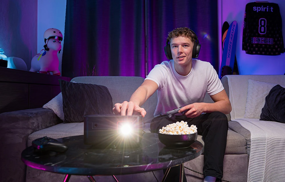 Un băiat care poartă căști ține un gamepad și se întinde spre proiectorul pornit așezat pe o masă cu popcorn