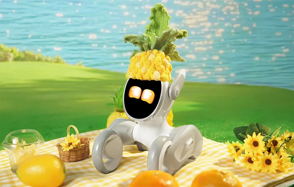 Robotul interactiv Loona într-un costum de ananas pe un covor de picnic înconjurat de flori și fructe, cu apă în fundal