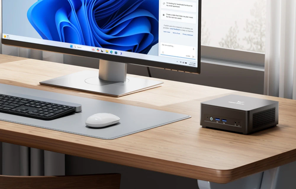 Un mini PC negru pe un birou din lemn, lângă un monitor cu Windows 11, o tastatură și un mouse, într-o cameră luminoasă.
