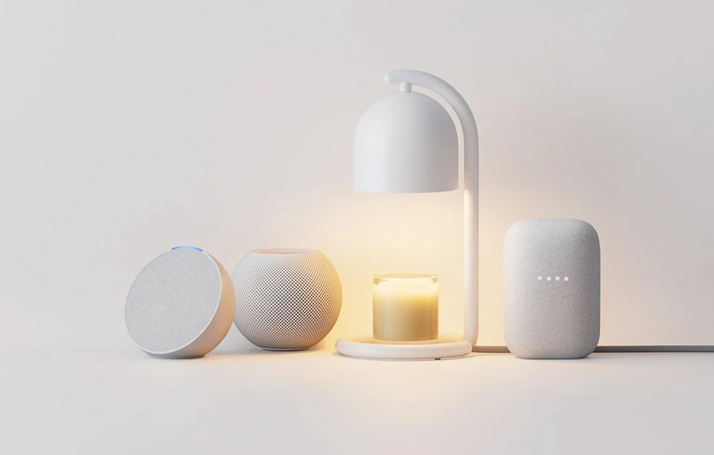 Un încălzitor de lumânări alb care luminează o lumânare parfumată din sticlă, așezat între trei difuzoare inteligente pentru casă: Amazon Echo Dot, Apple HomePod mini și Google Nest Audio, pe un fundal deschis.