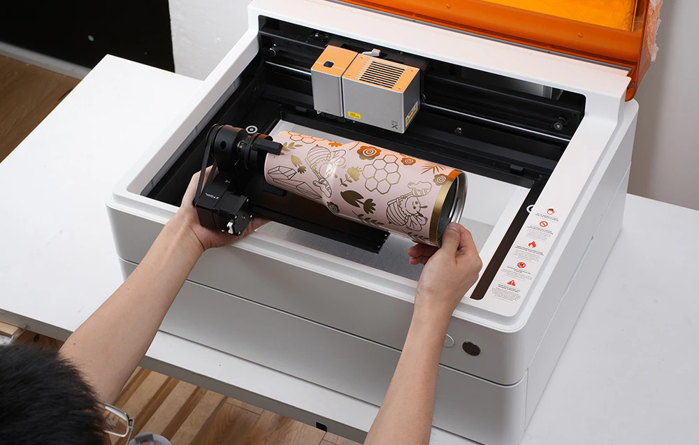 O persoană așezând un termos roz cu o gravură de albine în modulul rotativ din interiorul plotterului xTool deschis.