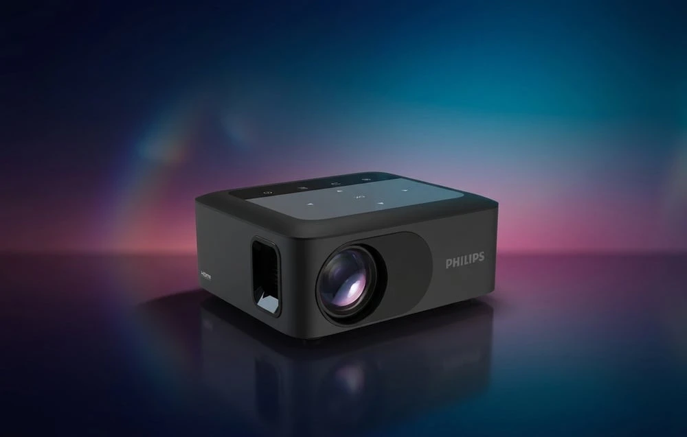Proiector PHILIPS negru cu inscripția „HDMI” pe lateral, așezat pe un fundal întunecat cu reflexii de lumină colorate