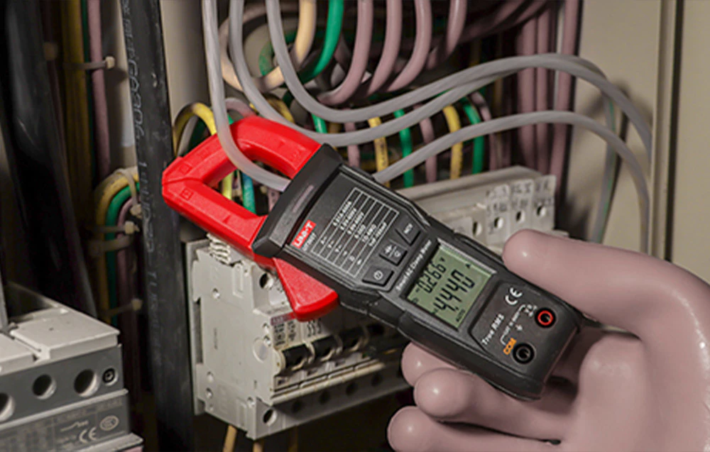 Un clește ampermetric UNI-T în mâna unui electrician care poartă mănuși de protecție, pe fundalul unor fire încâlcite și siguranțe.