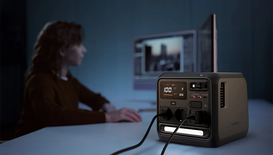 Stația de alimentare DJI Power 1000 Mini, cu două cabluri de alimentare conectate, se află pe un birou, iar în fundal o femeie stă și lucrează în fața monitorului unui computer