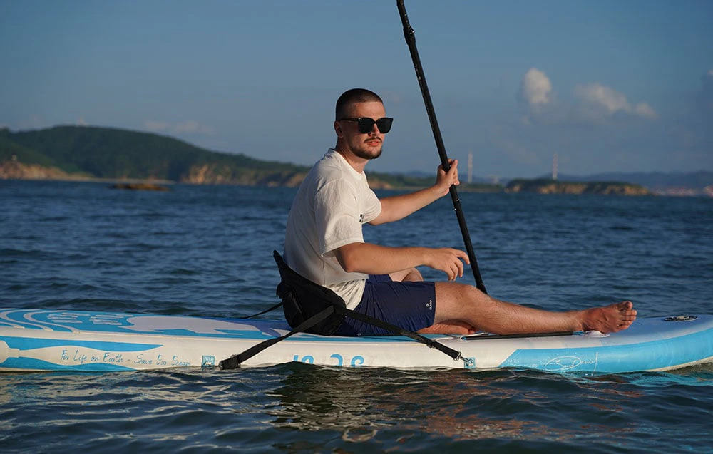 Un bărbat care poartă ochelari de soare stă pe o placă de surf cu scaun și ține o vâslă în mână