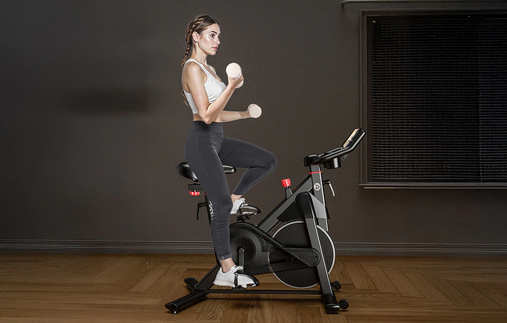 Persoană care se antrenează pe o bicicletă de spinning neagră, ținând în mâini gantere ușoare