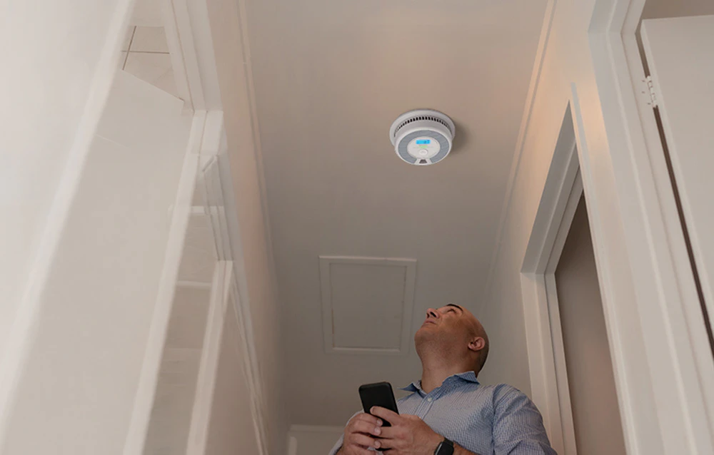 Un bărbat care stă în hol sub tavan se uită la senzorul inteligent de fum și monoxid de carbon instalat