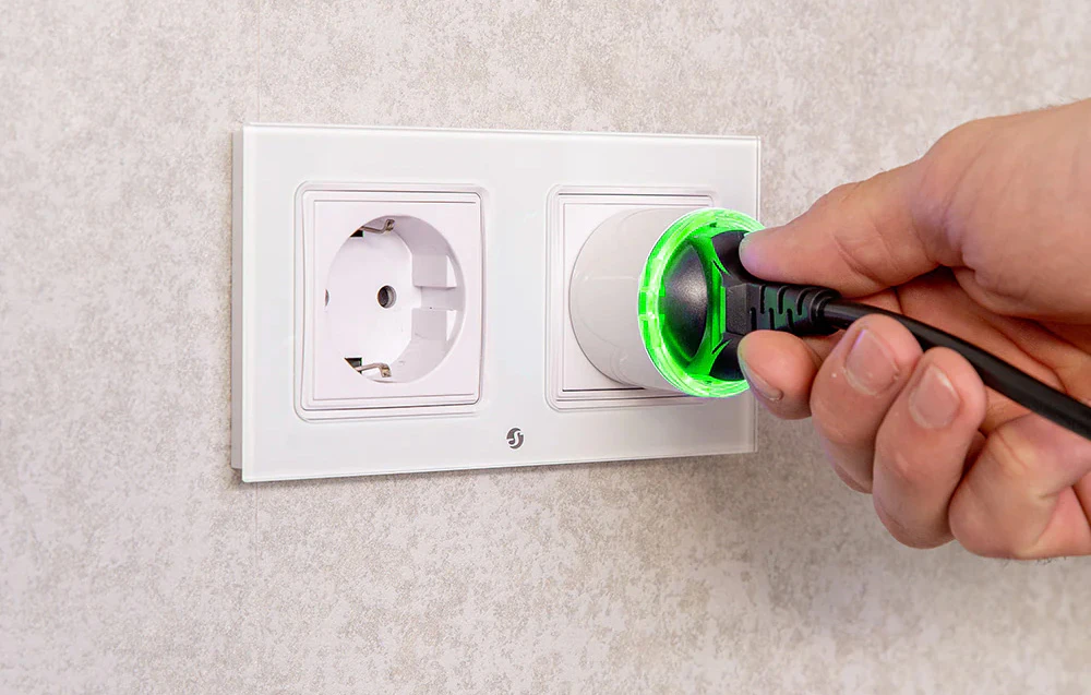 O mână introducând o priză neagră într-o priză inteligentă Shelly Plug cu iluminare LED verde, montată într-o priză de perete albă.