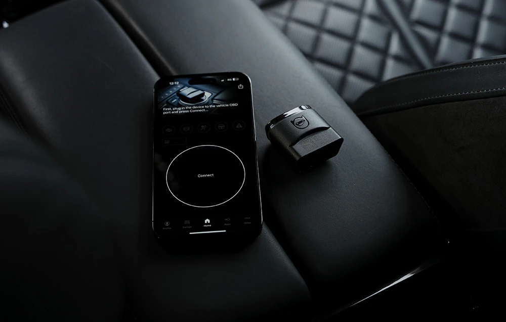 Smartphone cu aplicația Carly lângă adaptorul OBD2 pe un scaun auto negru matlasat