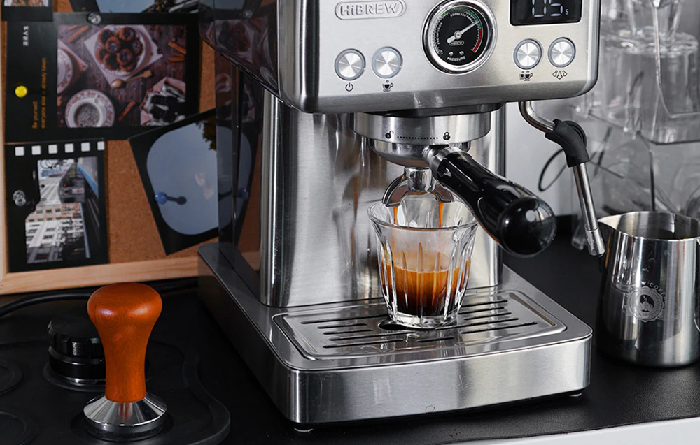 Aparat de espresso HiBREW într-o bucătărie de acasă, lângă accesorii pentru barista, cum ar fi un tamper din lemn și un ulcior metalic pentru lapte.