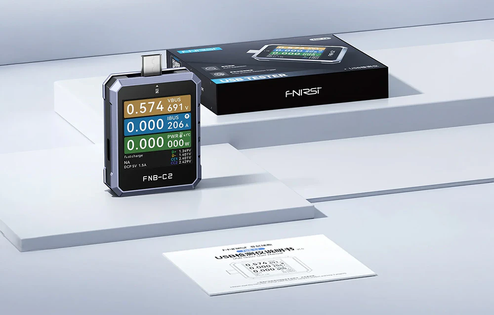 Un tester USB FNIRSI așezat lângă o cutie neagră și un manual de utilizare pe hârtie.