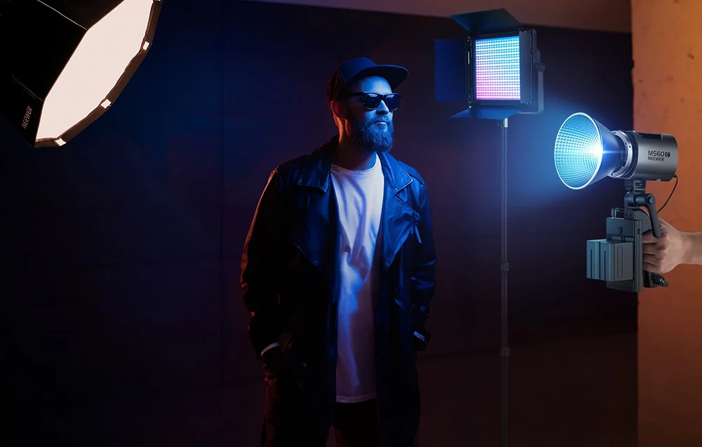 Un bărbat care poartă ochelari de soare și o jachetă pozează într-un studio foto iluminat de lumină colorată.