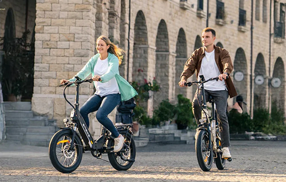 O femeie și un bărbat merg cu biciclete electrice printr-o piață înconjurată de clădiri din piatră