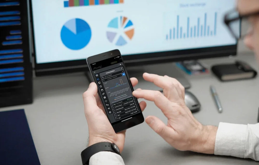 Un bărbat ține în mână un smartphone pe care se afișează un grafic al consumului de energie electrică; în fundal se vede ecranul unui computer cu diagrame.
