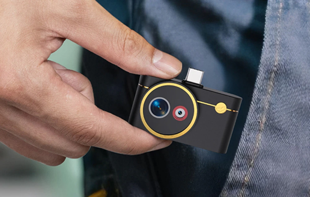 O mână scoate din buzunarul unei perechi de blugi o cameră termică mică, neagră, cu accente aurii și un conector USB-C