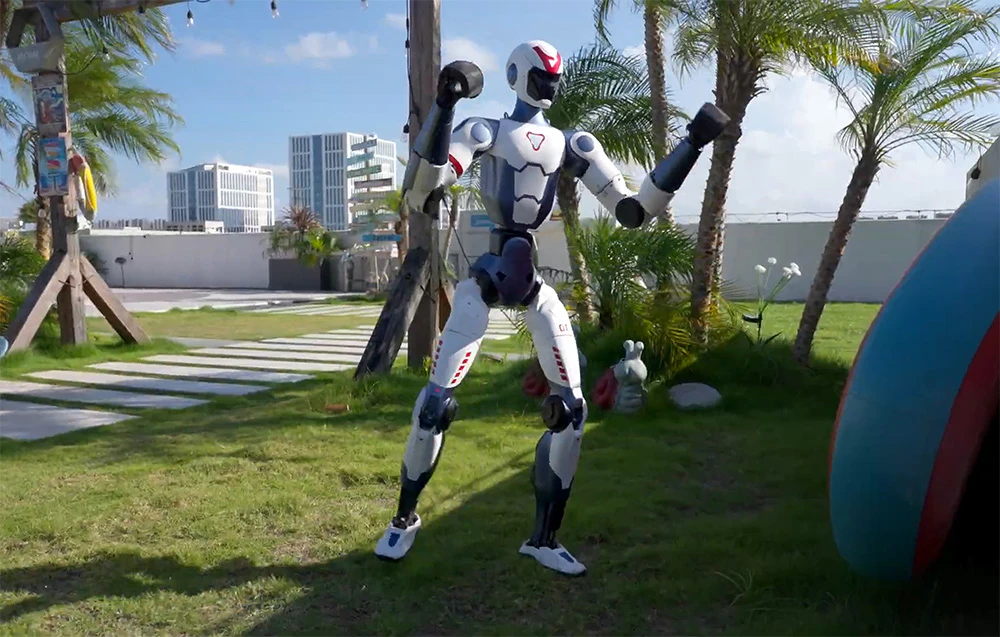 Un robot umanoid Unitree practicând arte marțiale pe iarba din fața unor palmieri