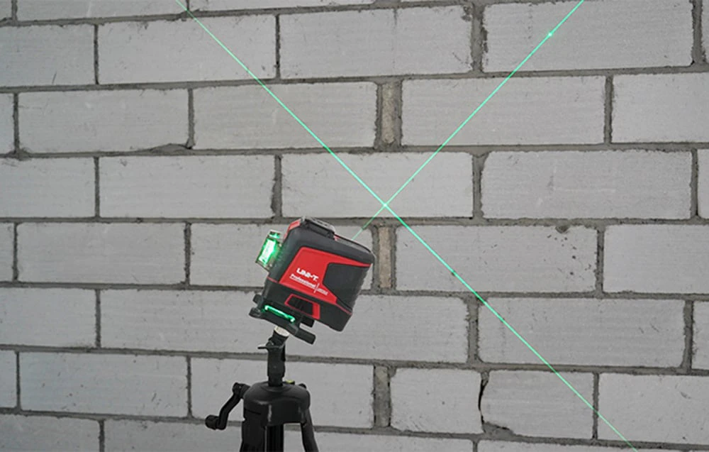 Nivelul laser montat pe un trepied proiectează două linii verzi care se intersectează pe un perete de cărămidă.