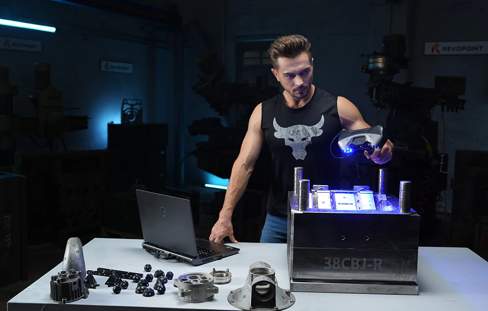 Un bărbat efectuează o scanare 3D a unei matrițe metalice, înconjurat de piese și un laptop pe o masă de lucru