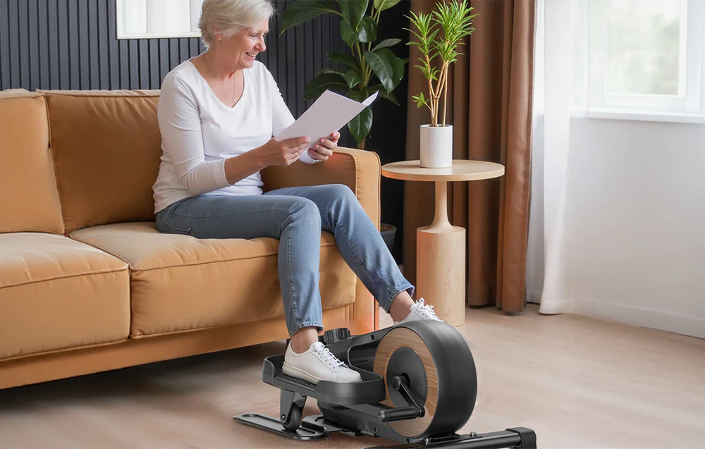 O femeie în vârstă stă pe canapea și face exerciții pe mini-eliptica Merach într-o sufragerie luminoasă.