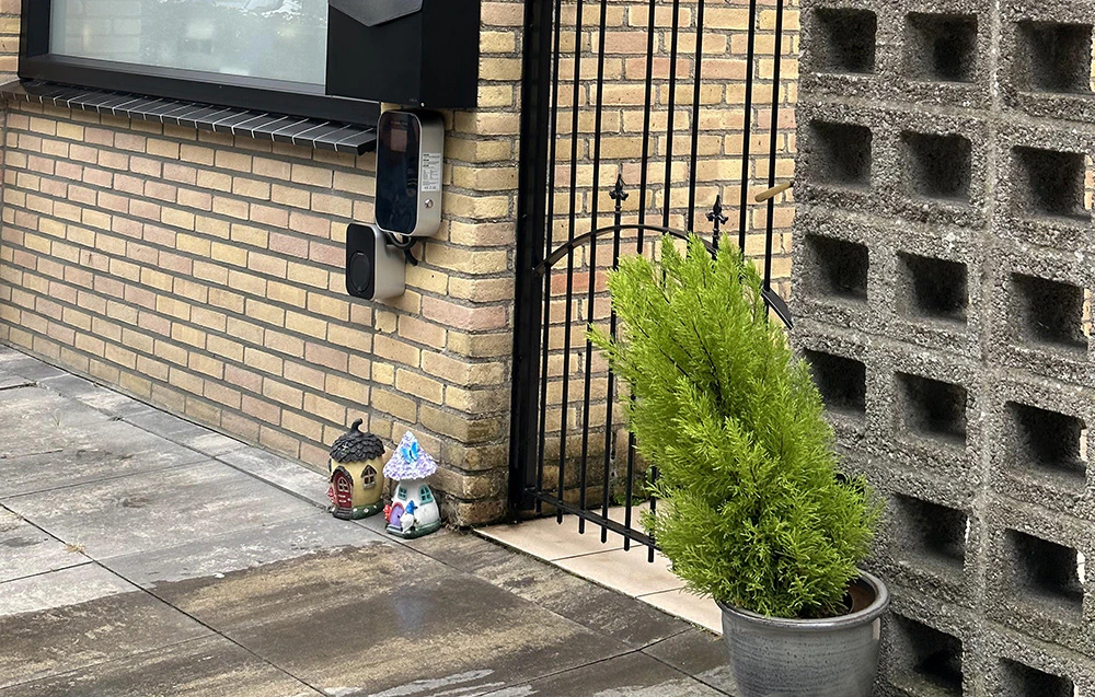 O stație de încărcare pentru mașini electrice montată pe peretele exterior al unei clădiri, lângă o poartă și o jardiniere cu o plantă.