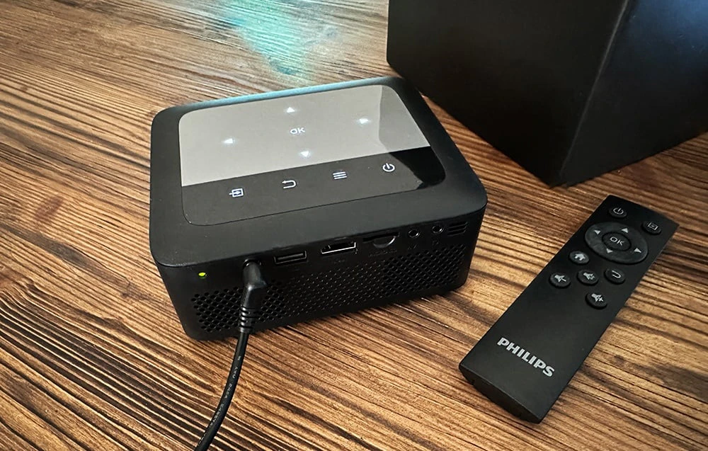 Vedere de sus a unui proiector negru cu un panou tactil iluminat și o telecomandă PHILIPS pe o masă de lemn