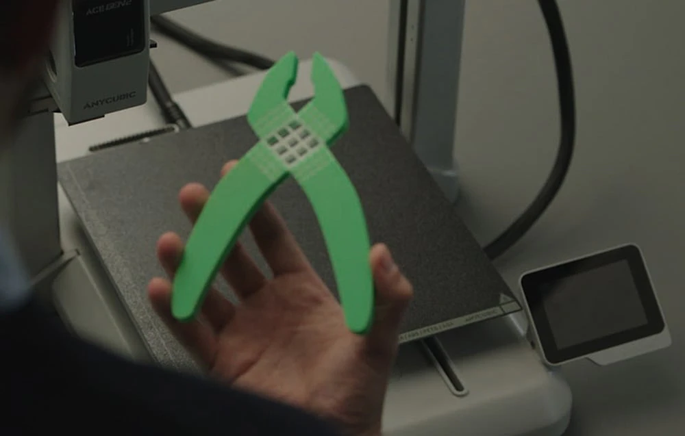 O mână ținând o clește verde, imprimată, pe fundalul mesei de lucru a unei imprimante 3D Anycubic